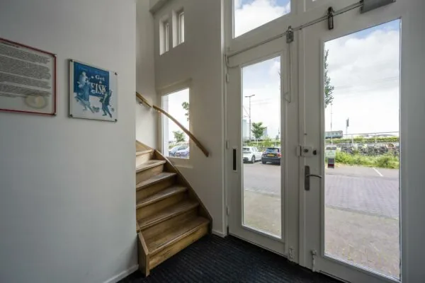 Entreehal met houten trap, glazen toegangsdeur en uitzicht op parkeerplaats aan de De Ruijterkade.